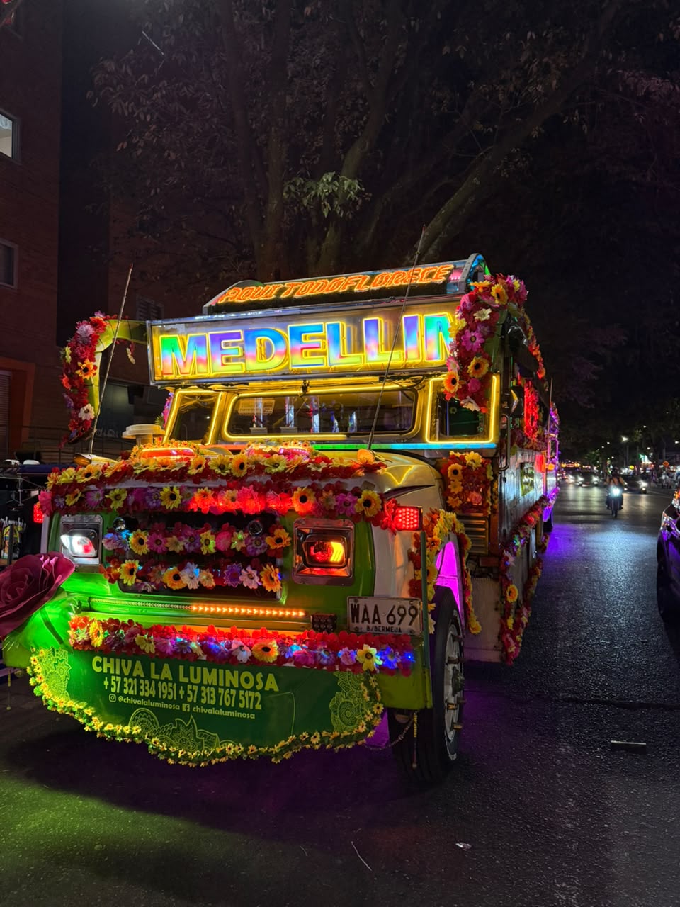 Chiva La Luminosa verde de noche, con el letrero de Medellín encendido y vestida de flores